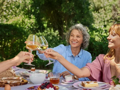 rose-garden-mothers-day Three people enjoying a cheerful outdoor brunch, toasting with wine glasses over a table filled with bread, fruits, and pastries. Lush greenery surrounds them.