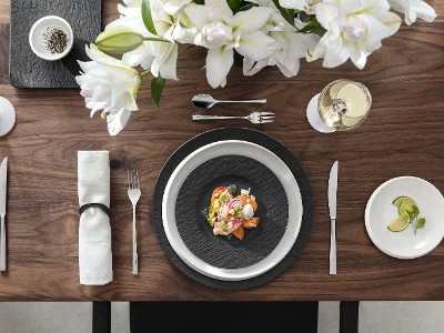Manufacture Rock & NewMoon – Modern Contrast, Pure Character Top-down view of a contemporary table setting combining Villeroy & Boch’s Manufacture Rock collection in matte black—featuring a gourmet plate, rectangular platter, and breakfast plate—with the minimalist NewMoon line in bright white, including a dinner plate, bread plate, and dip bowl. The contrasting shapes and surfaces create a striking yet harmonious design statement with handcrafted appeal.