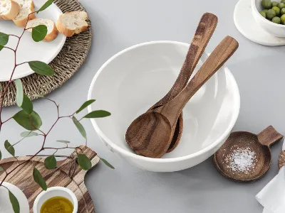 Artesano utensils in a white bowl, plus a small wooden bowl with salt, a chopping board with olive oil, a plate with bread, a bowl with olives and eucalyptus branches on a light-coloured surface from Villeroy & Boch.