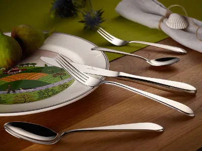 Sereno flatware set on Design Naif plates arranged on a wooden table. A white plate with a landscape motif, two figs, and a Sereno flatware set are arranged on a wooden table.
