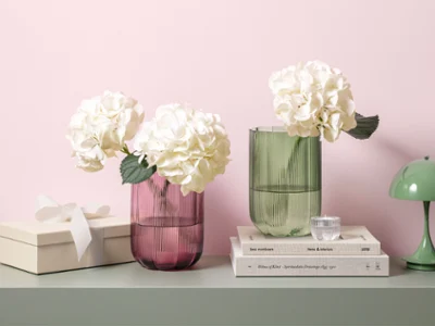Two Fleur glass vases with white hydrangeas on a table Two Fleur glass vases with white hydrangeas on a table next to stacked books, a gift box and a small green lamp.