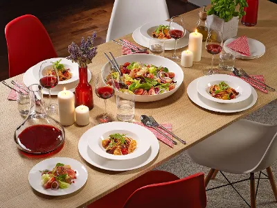 A dining table with For me plates full of salad, pasta, glasses of red wine, candles, and a carafe of red wine.
