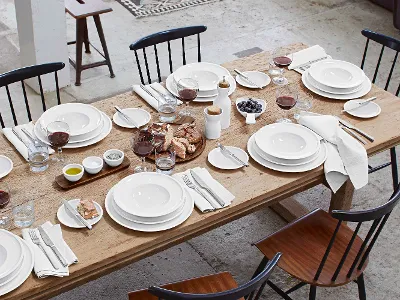 Stoviglie Artesano su un tavolo da pranzo in legno Un tavolo da pranzo in legno per sei persone con stoviglie bianche, bicchieri da vino rosso, tovaglioli, pane, uva, burro, sale, pepe, olio e bicchieri da acqua. I piatti e le posate sono disposti in ogni posto a sedere.