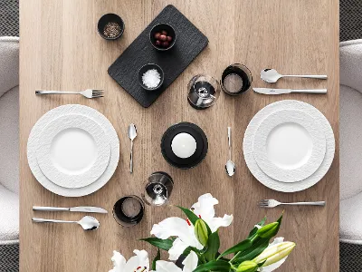 A beautifully set dining table with two white Manufacture plates, Piedmont flatware, glasses, a candle, a black tray with food, and a vase with white lilies on a wooden surface.