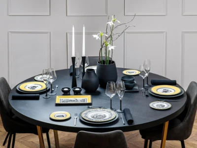 Elegant round dining table with black tableware, yellow-rimmed plates, white flowers in a vase, and lit candles, set in a modern room.