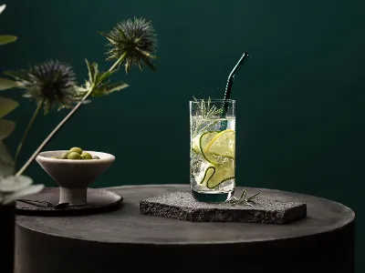 Bicchiere La Divina di Villeroy & Boch servito con stile Un bicchiere La Divina con fette di cetriolo e rosmarino, su una superficie di pietra con sfondo verde. Nel bicchiere c'è una cannuccia di vetro Artesano e accanto una ciotola di olive.