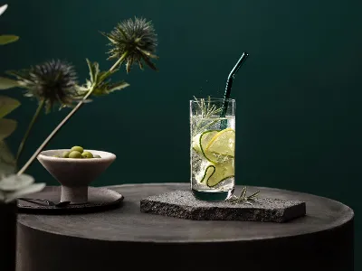 Stylishly served Villeroy & Boch La Divina glass A La Divina glass with cucumber slices and rosemary on a stone surface with a green background. An Artesano glass straw is in the drink and next to it is a bowl of olives.