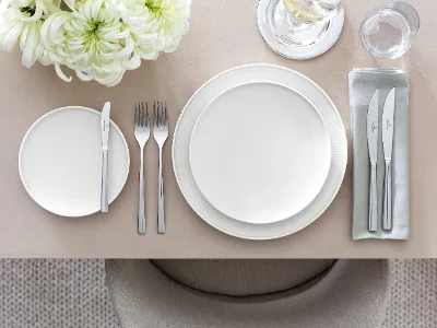 Table setting with Afina dinnerware and Piedmont flatware Sophisticated table arrangement with the Piemont collection from Villeroy & Boch, including a white dinner plate, a salad plate, a flatware set, glassware, and a floral centerpiece on a beige tablecloth.