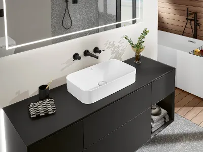 Lavabo Finion bianco su piano nero con miscelatore nero Un bagno moderno con un lavabo Finion rettangolare su un piano nero, uno specchio sopra di esso, rubinetti neri e una pianta in vaso. Sullo sfondo si vede una vasca da bagno.
