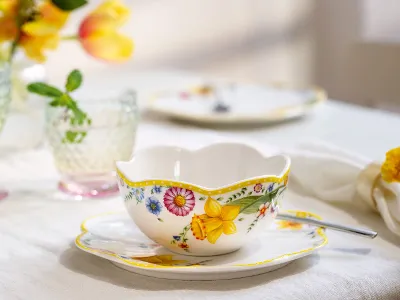 The ‘Spring Awakening’ teacup and saucer with spoon from Villeroy & Boch stand on the table next to crystal glasses and softly blurred flowers in the background.