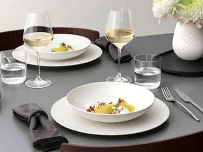A formal table setting with a Manufacture bowl on top of a plate A formal table setting with a white bowl on a white plate, glassware, and silver cutlery on a grey table and food.