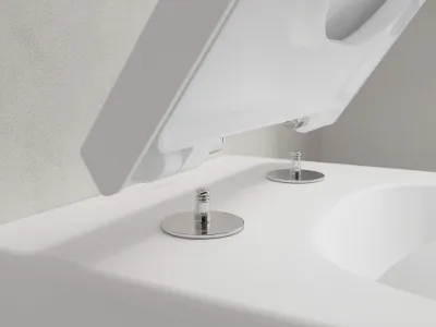Sedile per WC Venticello che si fissa alla toilette Primo piano di un WC bianco con coperchio aperto, che mostra le viti di fissaggio del sedile. Lo sfondo è una parete grigio chiaro.