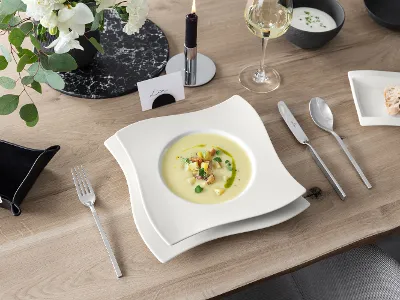 A bowl of creamy soup with garnish on a laid table with cutlery, wine, flowers and a small plate of bread.
