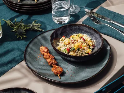 Summer treats on Lave crockery A plate of grilled chicken skewers and a bowl of mixed grain salad on a Villeroy & Boch Lave table setting, with glasses of water and floral decorations.