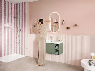 En kvinna sträcker sig framför spegeln vid tvättstället i ett stilfullt inrett pastellrosa badrum. Tvättstället består av ett vitt tvättställ med ett ljusgrönt underskåp och armaturen i krom från Skyla kollektionen. Den belysta, ovala More To See Lite+ spegeln har en praktisk hylla i vitt.