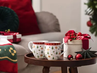 Two Christmas mugs, a gift box, and Christmas decorations on a small table next to a festively decorated living room.