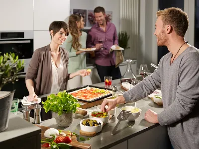 Villeroy & Boch dinnerware as a practical helper when baking pizzas Two people prepare pizza and fresh ingredients at a Kensington kitchen island from Villeroy & Boch. In the background, two other people are enjoying drinks and chatting.