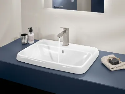 Lavabo Architectura bianco con rubinetto argento Un lavabo Architectura rettangolare bianco con acqua corrente, un rubinetto argentato, un asciugamano beige e un contenitore di sapone liquido su una mensola blu in un bagno.