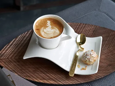 Golden NewWave spoon with matching cup and saucer A Villeroy & Boch NewWave latte cup with latte art on a wavy white saucer is combined with a small cookie and a golden spoon, all beautifully arranged on a wooden tray.