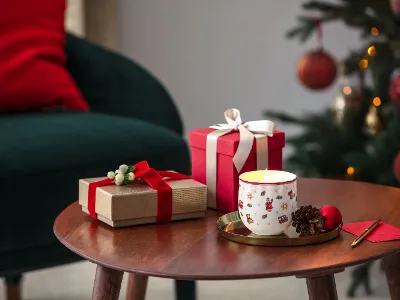 Toy`s Delight Candle as perfect gift A wooden table with two wrapped gifts, a lit candle in a festive mug, pinecones, and a red envelope near a decorated Christmas tree and a green chair.