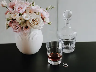 A Manufacture vase with pink and white flowers, a glass of whisky, a whisky carafe. A vase with pink and white flowers, a glass of whisky, a carafe and two rings on a black table.