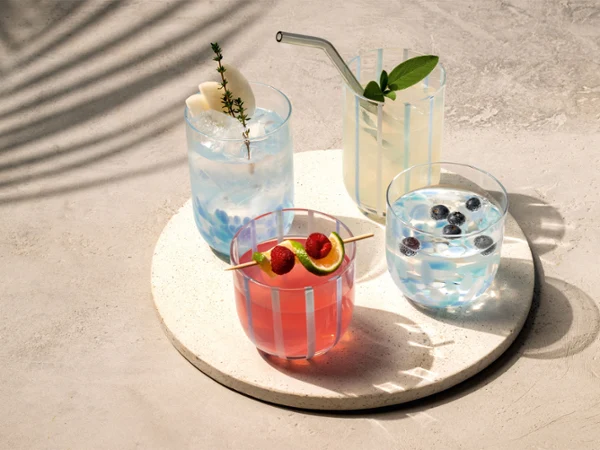 Quattro diverse bevande colorate in bicchieri Like Aqua con guarnizioni di frutta sono disposte su un vassoio bianco rotondo alla luce del sole.