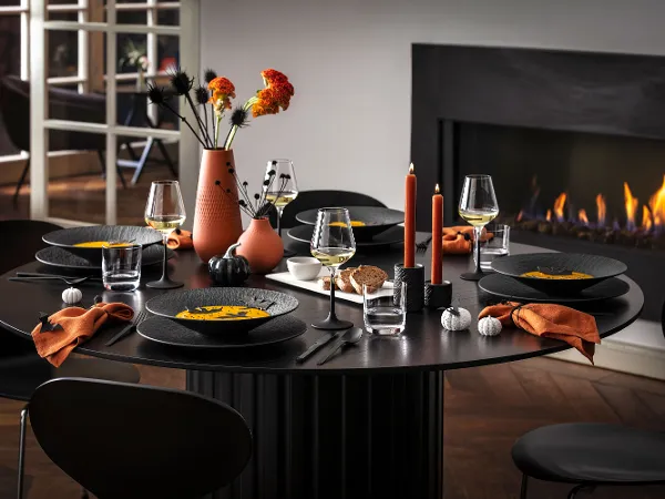 Zwart Manufacture servies met Halloween-decoratie Een moderne eettafel voor vijf personen met zwart Manufacture servies, oranje servetten en herfstdecoraties, waaronder vazen met bloemen en brandende kaarsen, naast een brandende open haard.