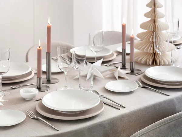De la vaisselle NewMoon sur une table de Noël dressée de façon moderne avec une décoration pastel Une table joliment dressée avec de la vaisselle blanche NewMoon, des verres à vin, des bougies roses, des couverts de table argentés et des décorations en papier sur une nappe beige. Un petit sapin de Noël en papier est également disponible.