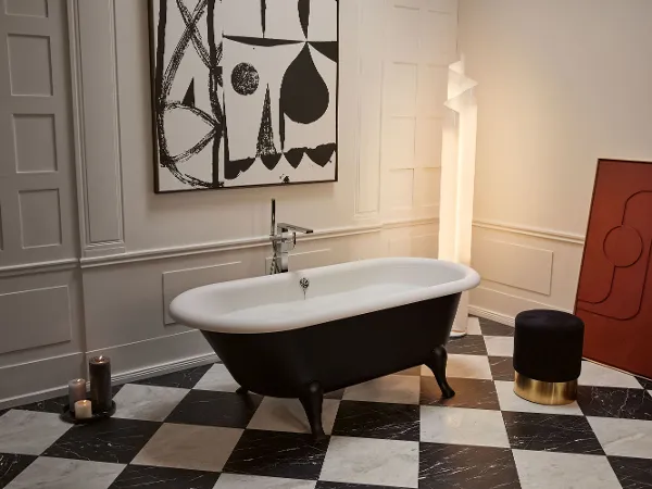 Freestanding black and white Hommage bath A stylish bathroom with a freestanding black and white Hommage bath, chequered floor tiles, a dark stool with a gold base, candles and a large geometric artwork.