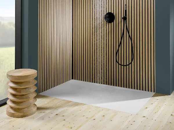 Shower tray from the Architectura bathroom collection in beautiful classic white Modern shower area with vertical wooden slatted walls, dark taps and a rectangular white plinth. On the left is a wooden stool and a large window offers a view outside.