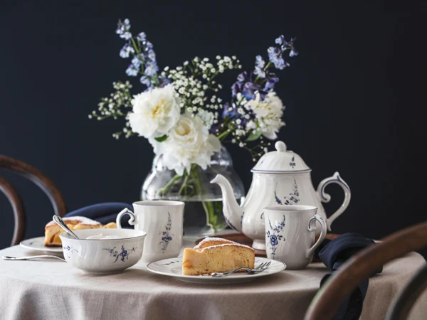 Vieux Luxembourg tablesetting with cake A round table set with vieuy luxembourg tea, teacups, cake slices, and a vase of white flowers against a dark background.