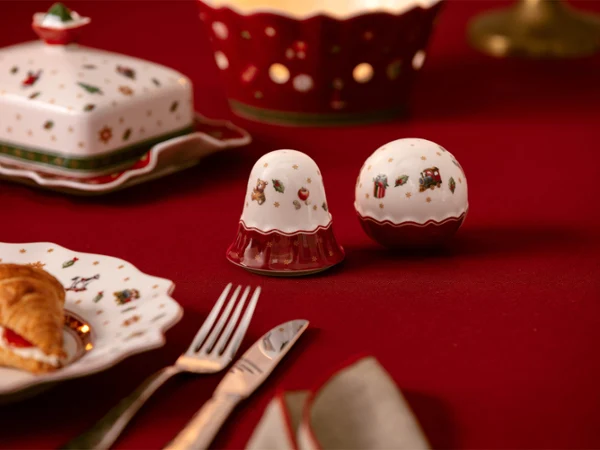 Salt and pepper shakers Saliere e pepiera con motivi natalizi su una tovaglia rossa, circondate da stoviglie festive e da un dolcetto.