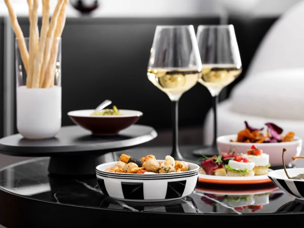Elegant cocktail and wine service featuring white wine, gourmet appetizers, breadsticks, and assorted snacks presented in modern black-and-white patterned bowls and plates on a marble table. The sophisticated setting creates a stylish atmosphere for entertaining and aperitivo-style gatherings.
