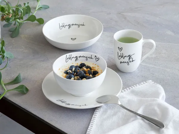 Statement Bow "Lieblingsmensch" gefüllt mit Blaubeeren Die Statement-Schüssel "Lieblingsmensch" mit Blaubeeren, dem passenden Teller sowie Henkelbecher und ein Löffel auf einem Tisch.
