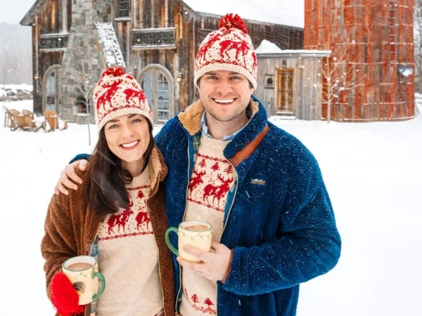 McCallister Sweaters  A smiling couple in matching winter hats and sweaters stands outside in the snow, holding mugs, with a rustic barn and silo in the background.
