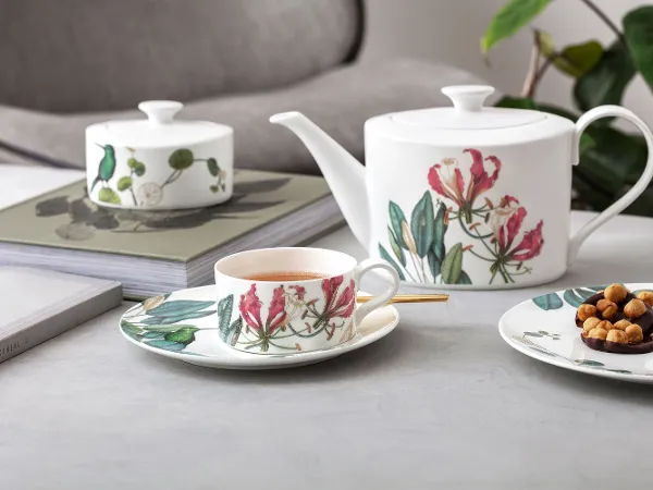 A teapot, a cup and a plate with snacks in a floral pattern are arranged on a table next to books and a plant in the background.