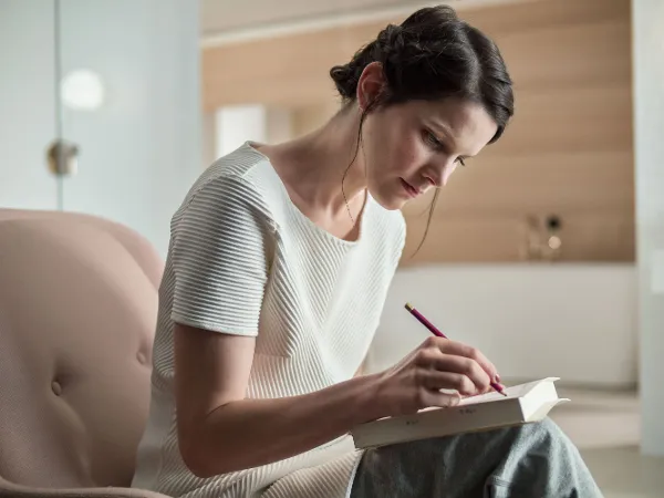 Gesa Hansen schreibt auf einem rosa Stuhl sitzend Eine Person mit dunklen Haaren und einem seitlichen Zopf sitzt auf einem rosa Stuhl und schreibt mit einem roten Stift in ein weißes Notizbuch. Sie trägt ein weißes Hemd und ist konzentriert.