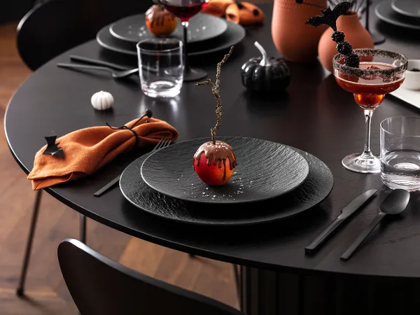 Zwart Manufacture bord met Halloween-decoratie en April met chocoladecoating Een zwarte eettafel is gedekt met zwarte Manufacture borden, oranje servetten en drankjes en versieringen met Halloween-thema, waaronder een appel in chocolade.