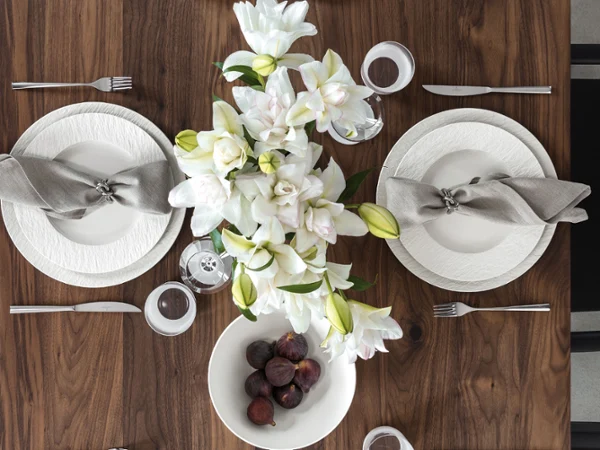 Festliche Tischdekoration Manufacture Rock Blanc mit weißen Tellern Vogelperspektive auf einen elegant gedeckten Walnussholztisch mit strukturierten weißen Manufacture Rock Blanc Porzellantellern, grauen Leinenservietten mit silbernen Ringen, einem natürlichen Centerpiece aus weißen Lilien und einer weißen Schale voller frischer, dunkler Feigen.