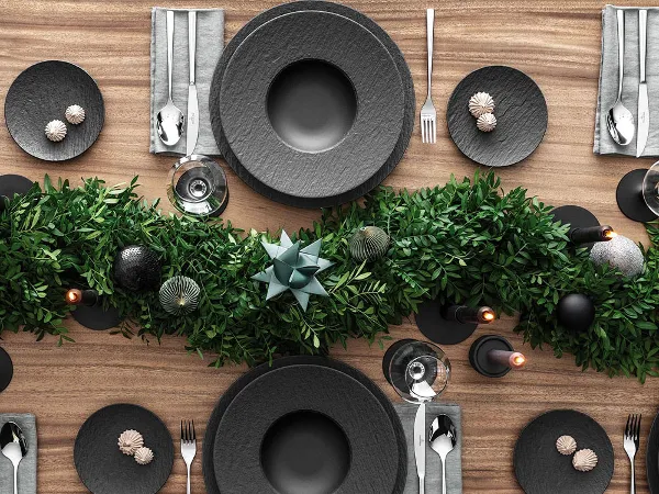 A top view of a festive dining table setting with black plates from the Manufacture collection. A top view of a Christmas dining table setting with black plates from the Villeroy & Boch Manufacture range together with Piemont cutlery. It is also decorated with a centrepiece of green foliage and candles on a wooden surface.