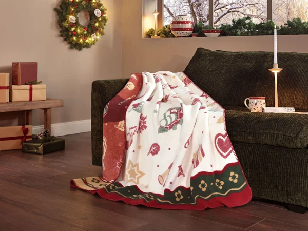 Toy's Delight blanket A cosy holiday-themed blanket lies over a dark green sofa in a festive living room with wrapped presents and decorations.