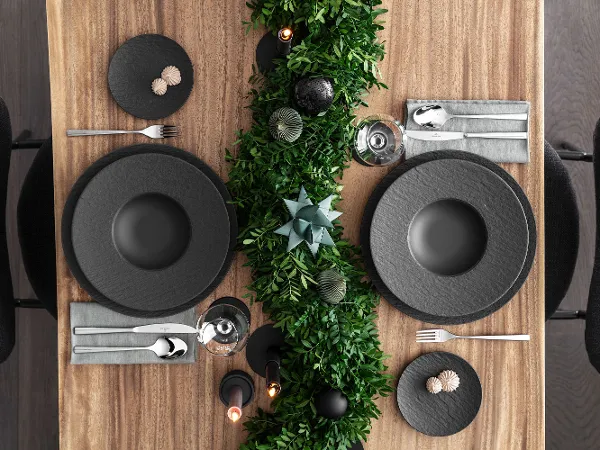 Platos negros Manufacture en una moderna mesa navideña Una bonita mesa de comedor con platos negros Manufacture, servilletas grises y cubiertos plateados. Una decoración de follaje verde recorre la mesa, acentuada por velas y objetos decorativos.