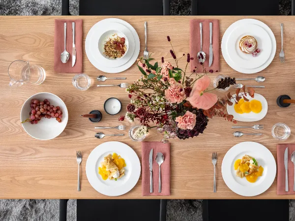 A rectangular wooden table is set for four people. There are plates with food, pink napkins, cutlery, candles, and a floral arrangement on the table. Grapes and spices are also present.
