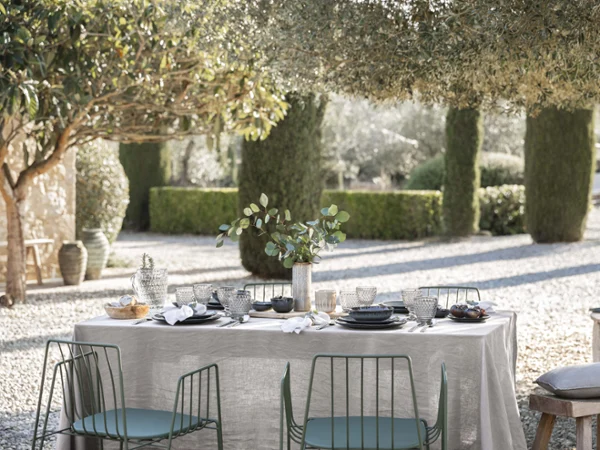 Outdoor dining area under trees with Lave plates on the table accompanied by elegant dark Boston glasses and Boston cutlery on a pebble surface.