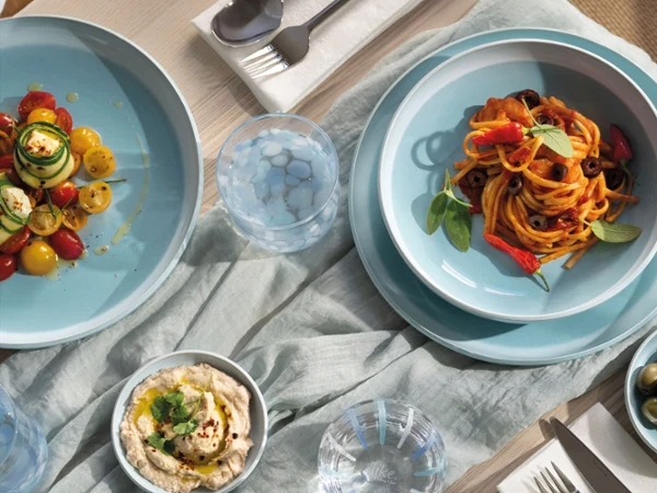 A table set with pasta on blue plates from the Perlemor Aqua collection A table set with pasta, salad, hummus, olives and utensils on blue plates from the Perlemor Aqua collection and a light-coloured tablecloth.