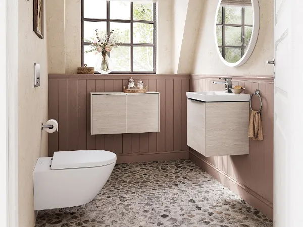 Gästebad mit Keramik und Möbeln der Villeroy & Boch Subway 3.0 Badkollektion Ein Badezimmer mit Fenster, Kieselboden, wandhängendem Waschbecken und Toilette. Über dem Waschbecken befindet sich ein runder Spiegel, an den unteren Wänden ist eine Holzvertäfelung angebracht, an den oberen Wänden hängen gerahmte Bilder.