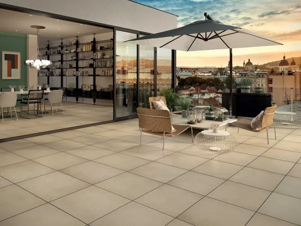Dachterrasse mit X-Plane Outdoor Fliesen Dachterrasse mit Korbstühlen, einem Couchtisch, einem Sonnenschirm und Blick auf die Stadt bei Sonnenuntergang. Der durch offene Glasschiebetüren sichtbare Innenbereich verfügt über einen Esstisch und ein Bücherregal.