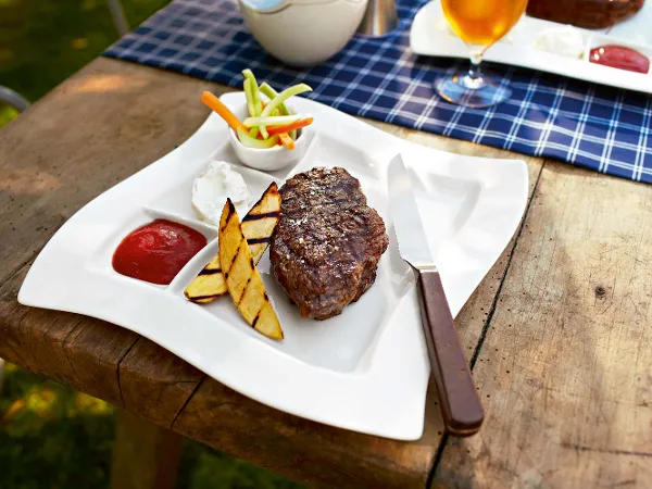 A grilled steak lies on a white NewWave grill plate with grilled potato slices, a dollop of sour cream, ketchup and sliced vegetables. A Texas steak knife rests on the edge of the plate. There is a drink in the background.