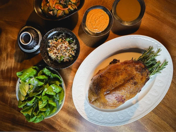 Overhead view of a roasted duck on a white platter with rosemary, surrounded by salad, potatoes, sauces, and a rustic wooden table.