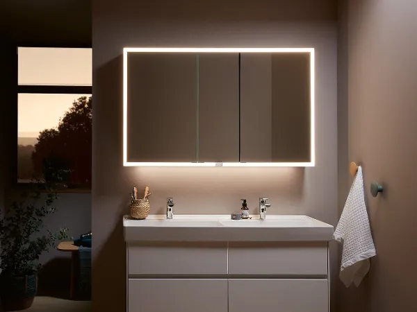 My View Now illuminated mirror with all-round LED lighting and a Villeroy & Boch washbasin A modern bathroom with a large illuminated mirror above a white washbasin and a vanity unit. A towel hangs on the wall and there are various toiletries on the shelf.